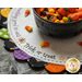 A decorative round table setting with a bowl of colorful candies in the center. The bowl contains various candy corn and festive treats. Surrounding the bowl is a white lace-like placemat with the words Trick or treat and playful Halloween-themed designs in orange, green, purple, and black.