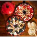 Two round quilted trivets with star patterns are displayed on a wooden surface. One trivet has a red, white, and gold design, while the other has a navy blue and white theme. A red cast iron pot and two wooden serving spoons are nearby. The image is branded with a logo in the corner.