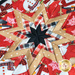 A close-up of a star-shaped quilt featuring a mix of festive fabrics in red, black, and cream colors. The design includes images of snowmen and various patterns like stripes and polka dots. The center is black, surrounded by pointed quilted sections.