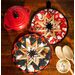 Two quilted pot holders are displayed on a wooden surface. One pot holder features a red and white snowman pattern, while the other is decorated in black and gold with a star design. A red pot and two wooden spoons are also visible in the image.