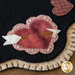 A close-up view of a decorative felt piece featuring a heart with an arrow through it, surrounded by smaller pink and red hearts, set against a black background with a beige stitched border.