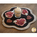 A decorative placemat with a scalloped edge featuring four pink hearts with arrows, embellished with beads, and a small glass candle holder at the center containing a lit candle. The background is black, contrasting with the heart designs.