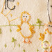 An embroidered image of a yellow chick with orange accents, surrounded by green stitching representing grass and small leaves. The background features letters and additional decorative elements.