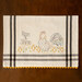 A decorative kitchen towel featuring a rooster and the phrase Rise & Shine along with small chick illustrations, bordered by gray stripes and a yellow scalloped edge.