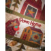 Cover of a quilt pattern book titled Down Home featuring colorful quilt blocks with house and star designs. Scissors and a wooden spool are placed at the bottom, along with a small red tomato pin cushion. The text One Sister is displayed at the bottom.