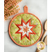 A round, quilted pot holder featuring a star pattern in orange, white, and green fabrics, set against a light background with wooden kitchen utensils and a floral arrangement in the upper left corner.