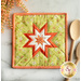 A decorative quilted hot pad featuring a star pattern in vibrant colors, including green, orange, and white, positioned on a light marble surface. Wooden cooking utensils sit beside it, and there's a hint of floral decoration in the background.