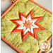 A colorful quilted pot holder featuring a star design with alternating orange, white, and green fabric. The star is surrounded by floral patterns on a light green background, bordered with a peach trim.