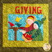 A quilted art piece featuring two stylized figures, one holding a basket and the other pointing, surrounded by bright yellow, green, and blue colors. The word GIVING is prominently displayed at the top in large red letters. There are birds depicted on a feeder beside them.