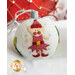 A decorative, felt Christmas ornament shaped like a white ball, featuring a playful elf character wearing a red hat and outfit. The ornament is embellished with colorful felt shapes and has a silver ribbon for hanging. The background includes soft, festive lighting.