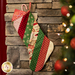 A colorful Christmas stocking hanging on a stone wall, adorned with red, green, and cream patterns, with a candy cane peeking out and a gift on top. A Christmas tree is softly blurred in the foreground.