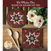 Two red hot pads with central folded star designs and red fabric resting on a wood table next to spoons and flowers.