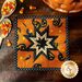 Orange Halloween themed hot pad with central folded star on wood table by jack-o-lantern, spoons, and candy corn.