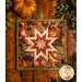 Square autumn themed floral hot pad with a central folded star design on wooden table next to small pumpkins.