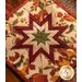 Square autumn themed floral hot pad with a central folded star design on wooden table next to small pumpkins.