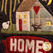 A quilted textile artwork featuring a colorful depiction of a house with a red door, a heart-shaped patch on the roof, and yellow windows. To the left, there are two animals: a sheep and a cow. At the bottom of the image, the word HOME is prominently displayed in large, textured letters.