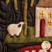 A decorative quilt featuring a small white sheep next to a green area with a tree, and a simplistic house with a red door and yellow windows. The background is dark, enhancing the colors of the fabric used in the design.