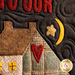 A close-up of a quilted textile featuring a house with a heart design and decorative elements including a star and crescent moon, accompanied by the text “Home Is Where Your Heart Is.”