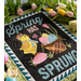 A colorful quilted decoration featuring a cheerful yellow chick wearing a hat, standing beside a basket filled with pastel-colored Easter eggs. The words Spring has Sprung are prominently displayed above and below the chick and basket. The background includes a green fabric with floral designs.