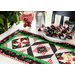 A table set with a colorful quilted table runner featuring triangular patterns in red, black, and green. There are napkins in a matching design and a bowl filled with apples, next to a jar covered with a checkered cloth. Lush green plants are visible in the background.