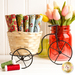 A decorative bicycle-shaped basket holds colorful fabric rolls, with a glass jar of tulips in the background. Spools of green and red thread are placed nearby.