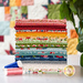 A stack of colored fabric rolls arranged neatly, showcasing various patterns and textures. At the bottom, there are two spools of thread—one blue and one pink. In the corner, there's a logo for a fabric brand. A patchwork quilt can be seen in the background.