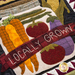 A close-up of a quilt featuring a design of vegetables, including orange carrots, red tomatoes, and purple eggplant, along with a banner that reads LOCALLY GROWN. The background includes a textured pattern resembling a jar and green leaves.