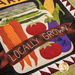 A quilted design featuring colorful vegetables including carrots, tomatoes, and eggplants, with the text LOCALLY GROWN prominently displayed on a banner across the center. The background has a textured pattern of various colors and shapes.