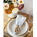 Flowers made of fabric being displayed in a white vase on small wood table next to table setting with cloth napkin.