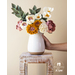 Flowers made of fabric being displayed in a white vase on small wood table.