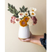 Flowers made of fabric being displayed in a white vase being held in outstretched hands.