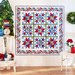 A colorful quilt with star patterns hangs on a white wall, surrounded by plants and decorative items.