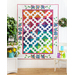 A colorful quilt featuring diamond and square patterns, displayed against a white wall with foliage.