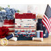 A neatly arranged stack of fabric squares in red, white, blue, and cream colors, featuring various patterns. Spools of thread in coordinating colors are placed in front, alongside a small American flag and decorative flowers.