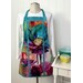 A colorful floral apron displayed on a mannequin against a white wooden backdrop, with kitchen utensils nearby.