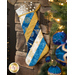 A decorative holiday stocking hangs from a stone fireplace, featuring blue, white, and gold stripes with snowflake designs. A silver snowflake ornament decorates the top of the stocking. Christmas ornaments are visible in the surrounding greenery.