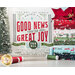 A festive display featuring a decorative wall sign that reads I Bring You Good News of Great Joy That Will Be for All the People. For unto you is born this day in the city of David a Savior who is Christ the Lord. The background includes a small evergreen tree and neatly stacked rolls of holiday-themed fabric in red, green, and white patterns.