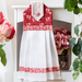 White hanging towel made with red and pink floral fabrics hanging from a white shelf with pink roses behind on a shelf, and leaves and pink tulips in the foreground
