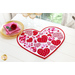 Heart shaped table topper with pink and red hearts in different sizes and prints filling the center on a white table with a gold rimmed plate, pink napkins, a red rose, and gold wine glasses in front of a window
