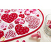 Close up angled shot of heart table topper with different sized hearts in red and pink on a white background on a white table with a dish of red and pink spools of thread and buttons scattered on the table