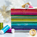 A neatly stacked array of colorful fabric pieces in various shades, including pink, green, and blue, with spools of thread in complementary colors arranged beside them. The setting has a bright, festive feel.