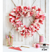 Heart shaped scrappy wreath made up of pink, red, and white fabric strips hanging on a white wooden wall with cross beams and crafting supplies and a pen holder on the white countertop in the foreground.