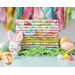 A stack of colorful fabrics with Easter-themed patterns sits on a table surrounded by pastel-colored Easter eggs and green grass-like filling. A small plush bunny with pink ears is in front of the fabric.