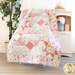 Image of Comfort of Psalms quilt made with pink, blue, and cream floral fabrics draped over a small chair with a small white shelf behind it with pink flowers and books on it with a houseplant on the opposite side.