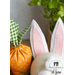 Close up of the tops of fabric carrots and the top of the a white felt bunny in front of a grey wall with a small houseplant in the background