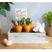 A white washed planter box filled with green moss and fabric carrots with a bunny figure diving into the box and a sign that says 