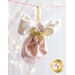 A decorative fabric ornament shaped like an angel, featuring a white bow and a pink polka-dotted body, adorned with a gold ribbon and charm, hanging from a string with blurred lights in the background.