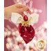 A person holding a handmade ornament shaped like an angel, featuring a red fabric body decorated with white floral patterns and a white bow at the top. The ornament has a gold ribbon accent. The background is softly lit with blurred lights.