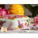 Complete side view image of a floral gold, pink, and mint pincushion with a dozen pins stuck in it, set on a white table with buttons, gold scissors, pink flowers, and a yellow honey pot behind it