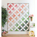 A colorful quilt featuring a geometric pattern of crosses in shades of pink, green, and white hangs on a wall, surrounded by potted plants and decorative items.